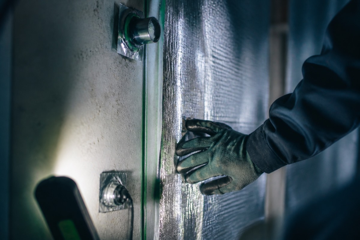 A gloved hand sealing a mold-resistant moisture barrier over a concrete wall under a bright inspection light.